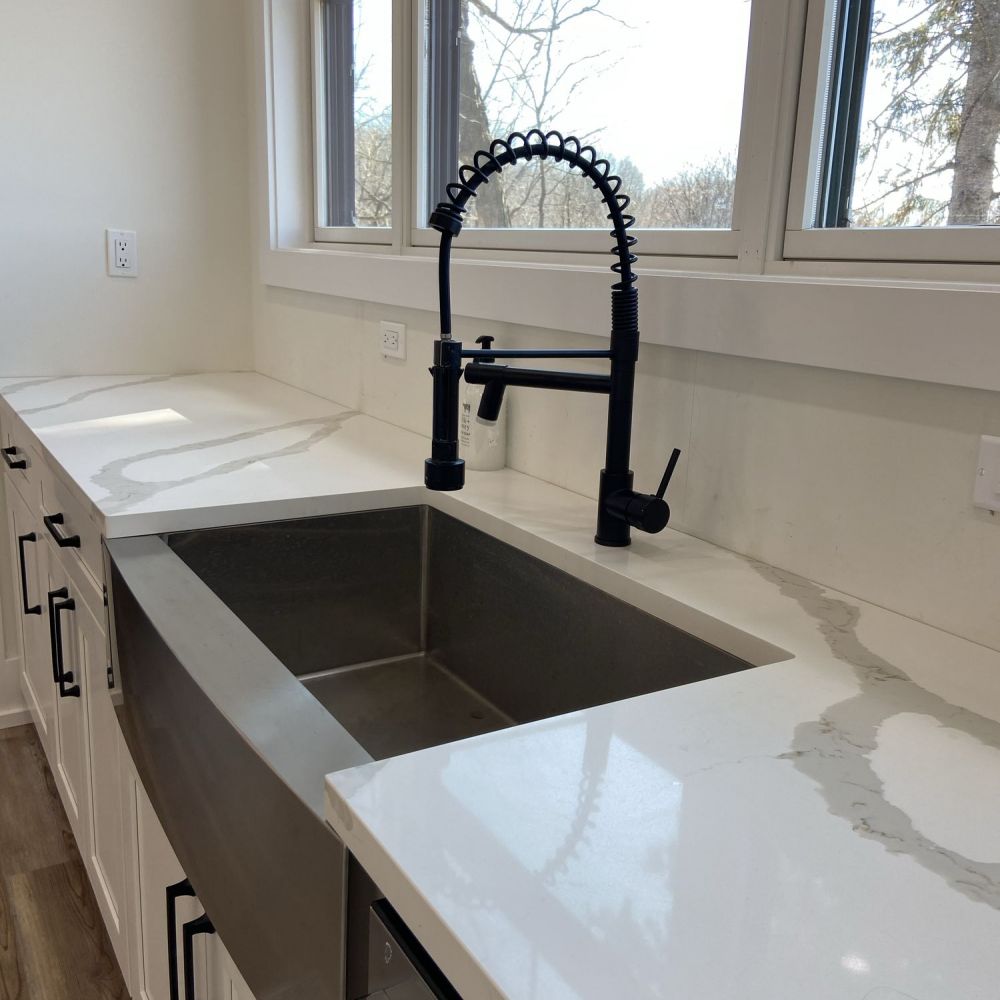Modern kitchen sink with black pull-down faucet, white countertops, and large windows overlooking trees.