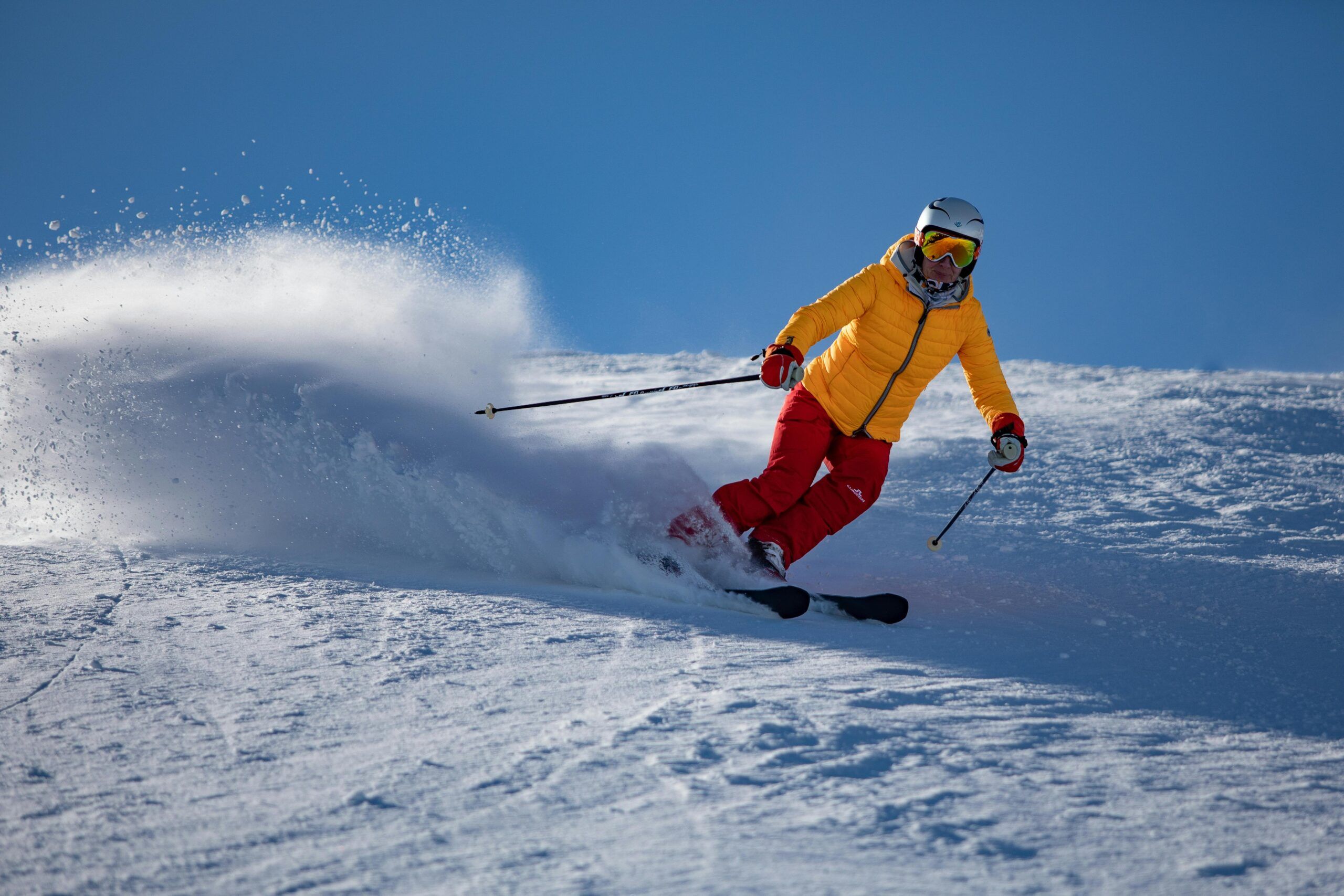 Person skiing downhill in bright sunlight, wearing a yellow jacket and red pants, creating a spray of snow at Mountain Crest Reserve.