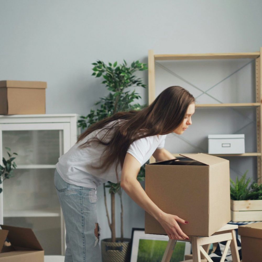Woman moving a cardboard box in a room with unpacked items, plants, and shelves for her new Mountain Crest Home.