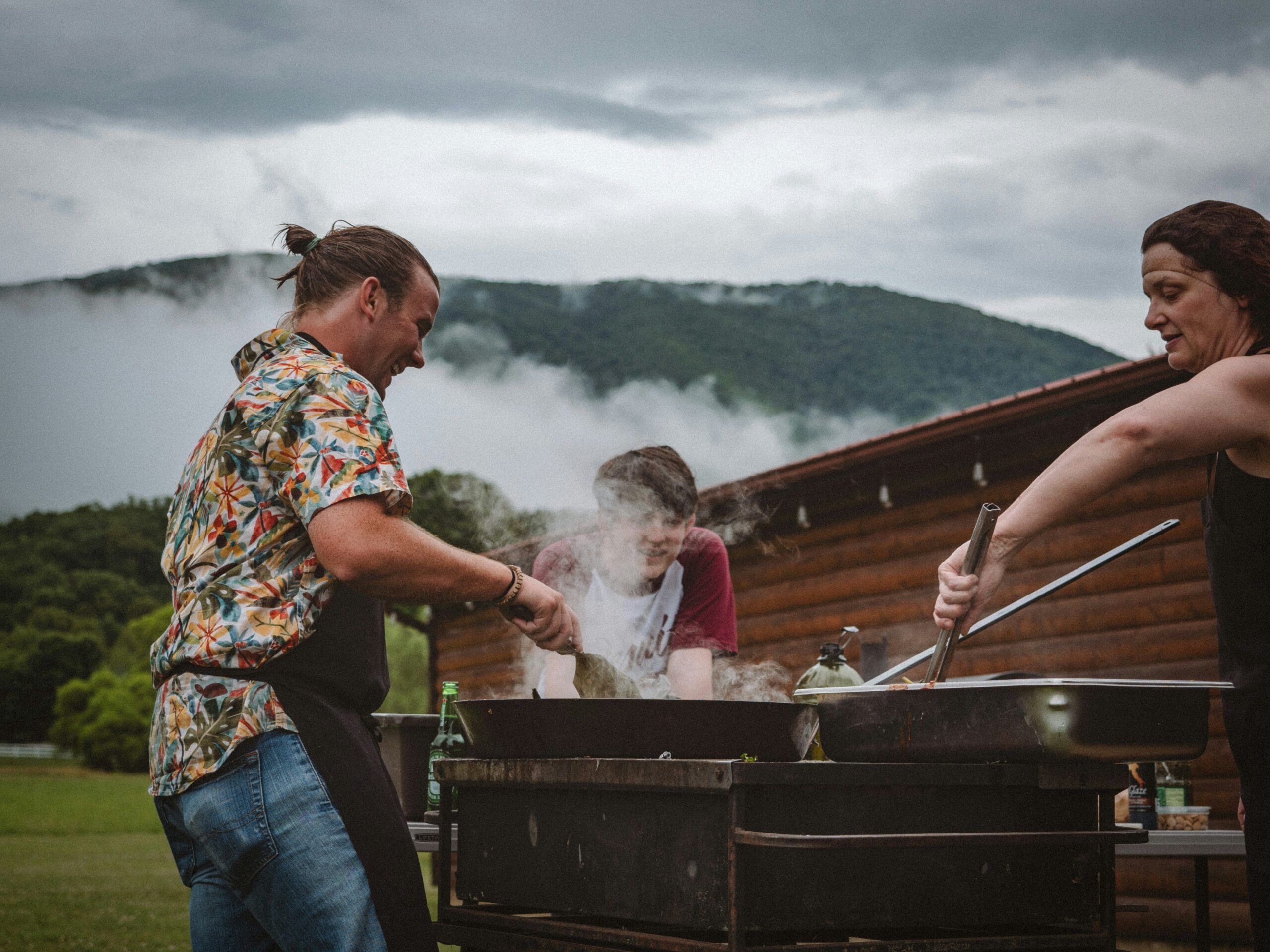 Three people cooking at a large grill outdoors, surrounded by mountains and mist at Mountain Crest Reserve.