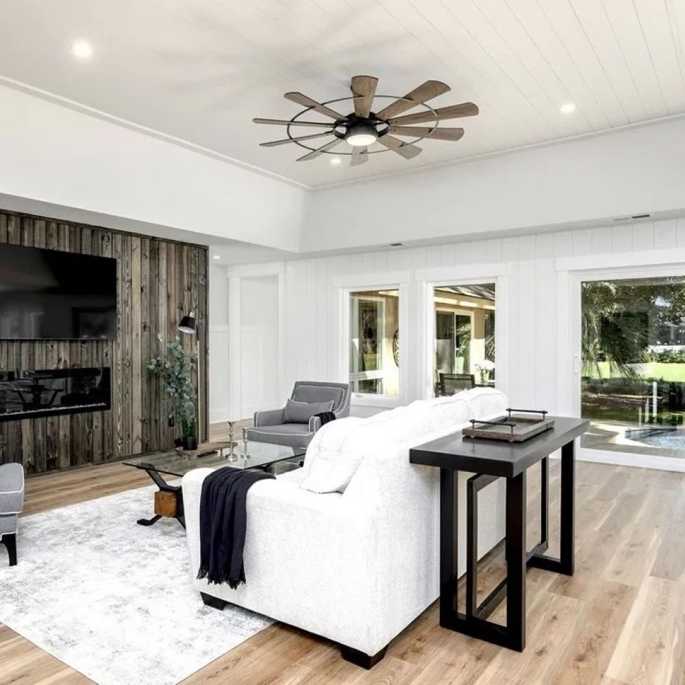 Modern living room with white walls, large windows, a gray couch, armchairs, wood accent wall, and ceiling fan at Mountain Crest Reserve.