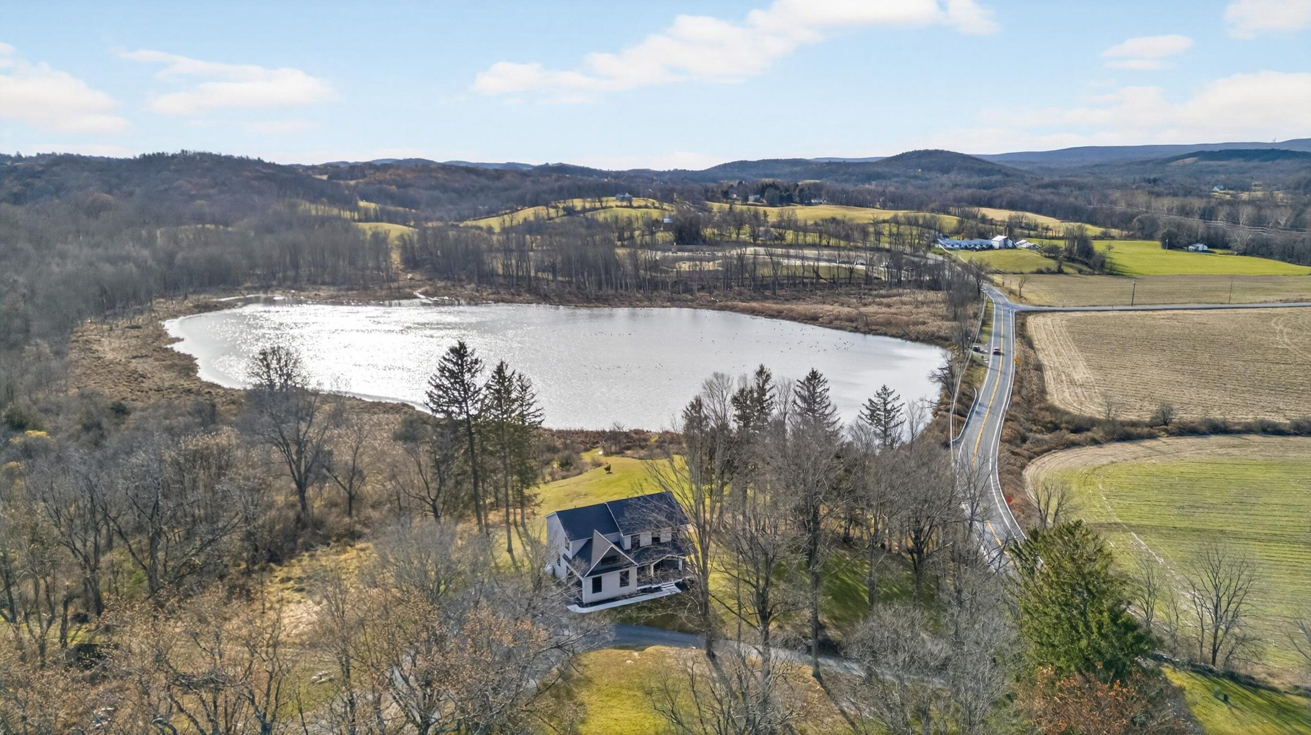 A house sits near a lake surrounded by trees and rolling hills under a partly cloudy sky.
