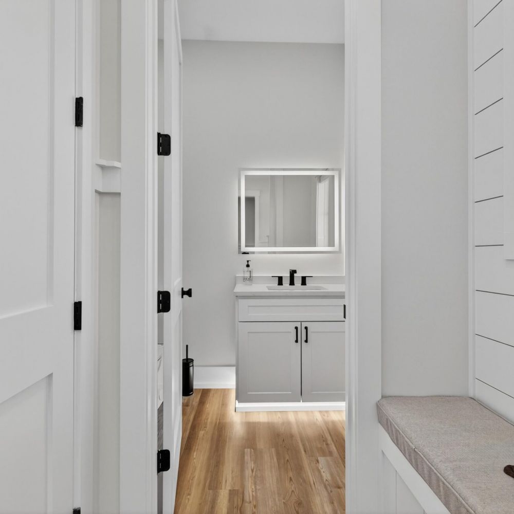 Minimalist bathroom with white walls, light wood floor, and a vanity with a mirror and black fixtures.