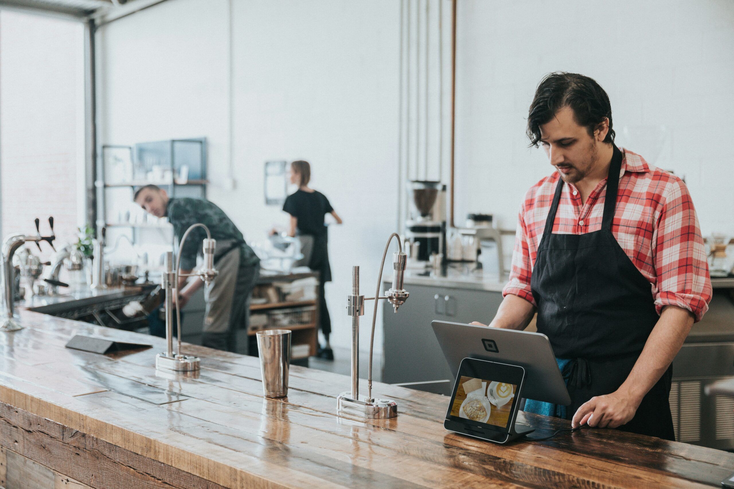 Barista in red plaid shirt and black apron using a tablet at a coffee shop counter with another person in the background at Mountain Crest Reserve.