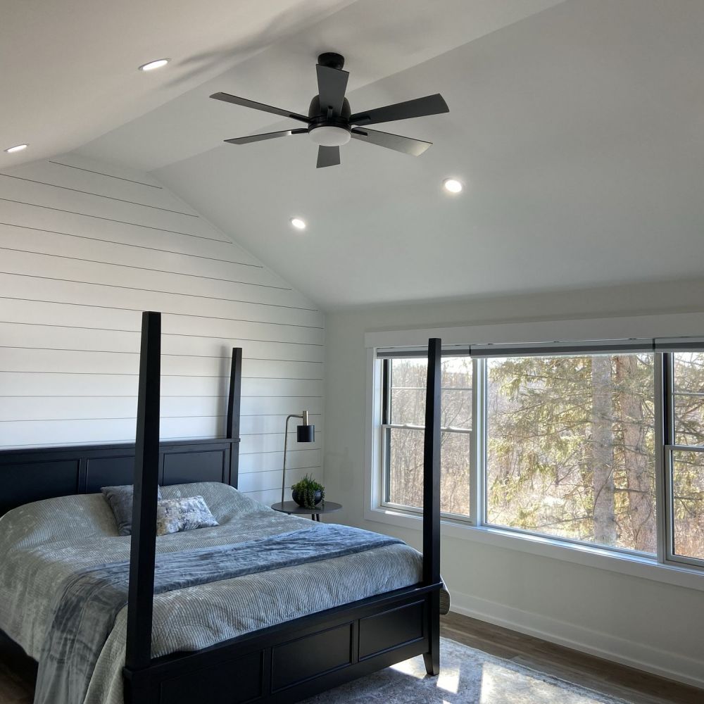 A bedroom with a black four-poster bed, shiplap walls, ceiling fan, and large window overlooking trees.