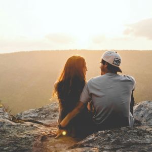 A couple sits on a rocky cliff at sunset, embracing as they look at the distant hills under a glowing sky.