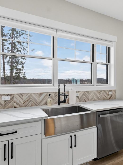 Modern kitchen with a large window, farmhouse sink, white cabinets, and a view of trees and hills outside.